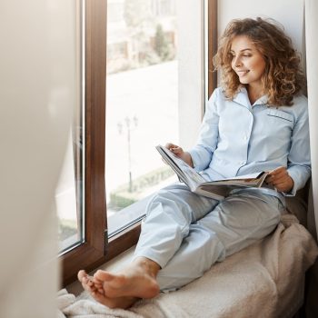 Charming businesswoman having relaxing time at home. Pleased good-looking adult woman in nightwear sitting on window sill and gazing at street, holding fashion magazine, reading about lifestyle. Copy space