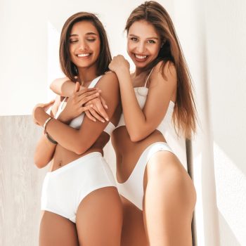Portrait of two young beautiful smiling women in white lingerie. Sexy carefree models posing at the balcony in the early morning. Models hugging and enjoying their morning